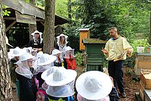 Kinder stehen mit Imkerhüten vor einem Imker, der Bienenwaben in der Hand hält.