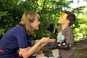 Eine Frau lacht mit einem Jungen, der Handschuhe und Ellenbogenschoner trägt sowie eine gelbe Folie vor seinen Augen hat.