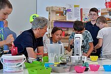 Kinder sitzen mit einem Erwachsenen an einem Tisch mit einem Mikroskop und bunten Behältern