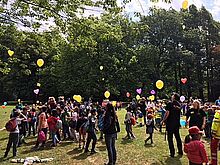 AUf einer Wiese lassen viele Kinder und Erwachsene Luftballons mit angebundenen Nachrichten steigen