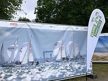 Eine Beachflag der Stiftung "Haus der kleinen Forscher" vor einem großen Plakat mit Segelbooten drauf