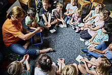 Kinder sitzen mit einer im Kreis auf dem Boden und halten ihre Hände geöffnet. Sie warten darauf, dass die Frau ihre Hände mit Erde aus einer Box füllt., sodass