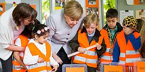 Angela Merkel lässt sich von Kindern in oranger Warnweste etwas zeigen