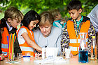 Bundeskanzlerin Angela Merkel forscht gemeinsam mit Kindern in orangen Warnwesten an einem Tisch