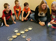 Eine Erzieherin sitzt mit Kindern auf dem Boden und untersucht verschiedene kleine Gegenständi in Glasschüsseln